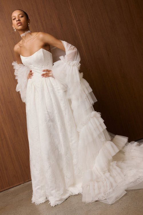 A model wearing a strapless white wedding gown with dramatic layered tulle sleeves and a long ruffled train, posed against a dark wood-paneled wall.