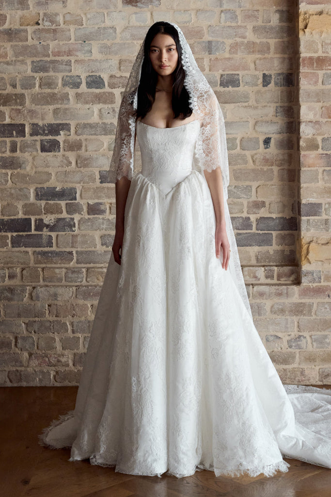 A woman wearing a white lace wedding gown with a matching lace veil stands in front of a rustic brick wall.
