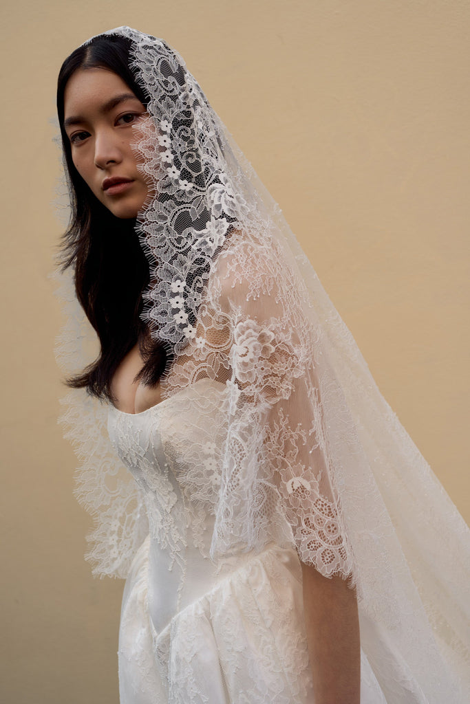 A woman wearing a white lace bridal veil and a white wedding dress, photographed against a plain warm beige studio background.