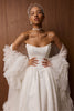 A woman modeling a strapless white wedding gown with dramatic ruffled sleeves, a sheer veil headpiece, and a jeweled choker against a warm wood-paneled background.
