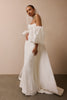 A woman wearing an elegant off-the-shoulder white wedding gown with dramatic puffed sleeves, a long train, and a sheer veil, standing against a warm beige wall.