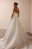 A woman wearing a strapless white wedding gown with a structured bodice and a full, flowing train, photographed in a softly lit indoor setting with a plain beige wall_XS