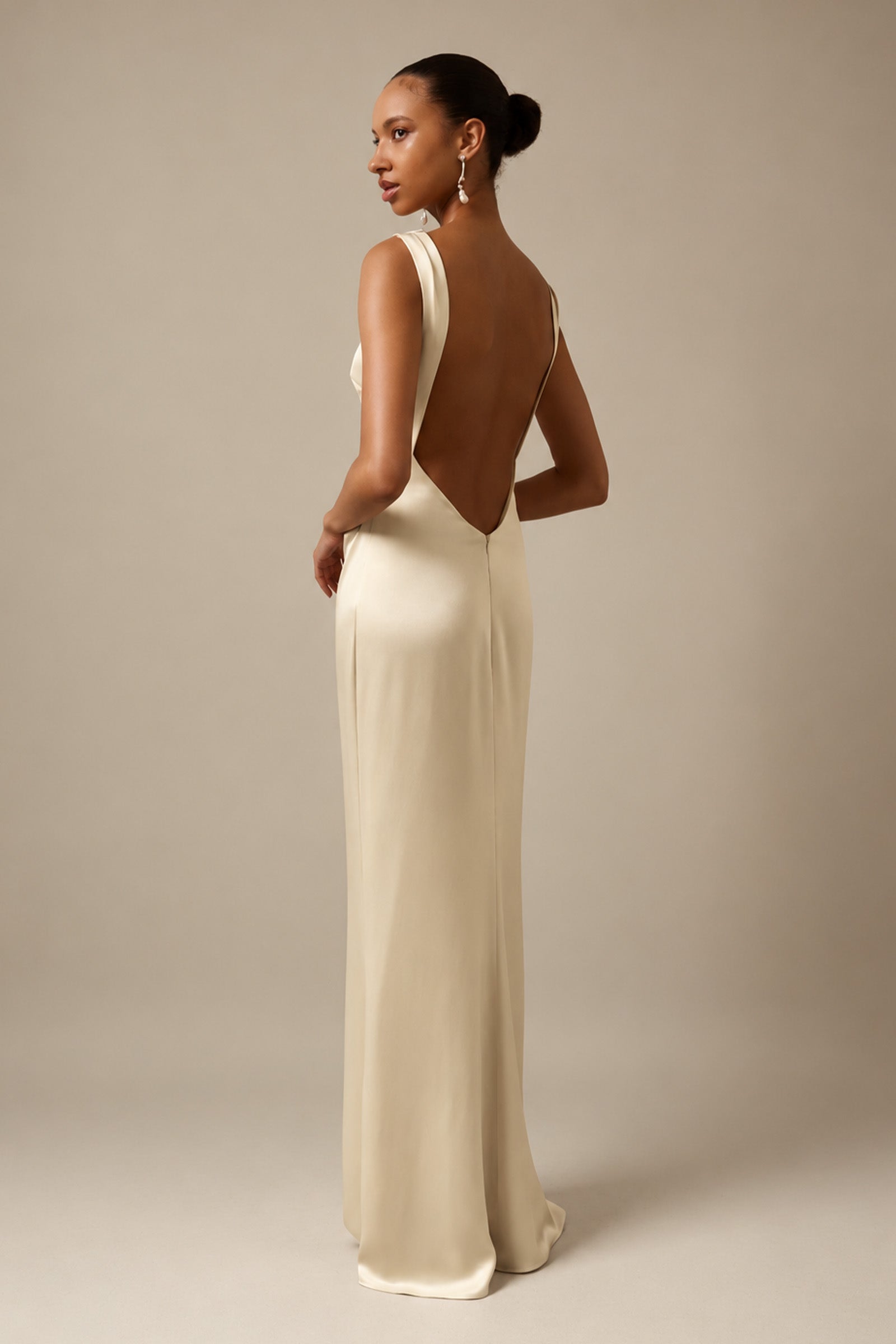 A woman modeling a floor-length cream satin gown with a deep open back, shown from a rear three-quarter angle against a warm beige studio backdrop_XS_