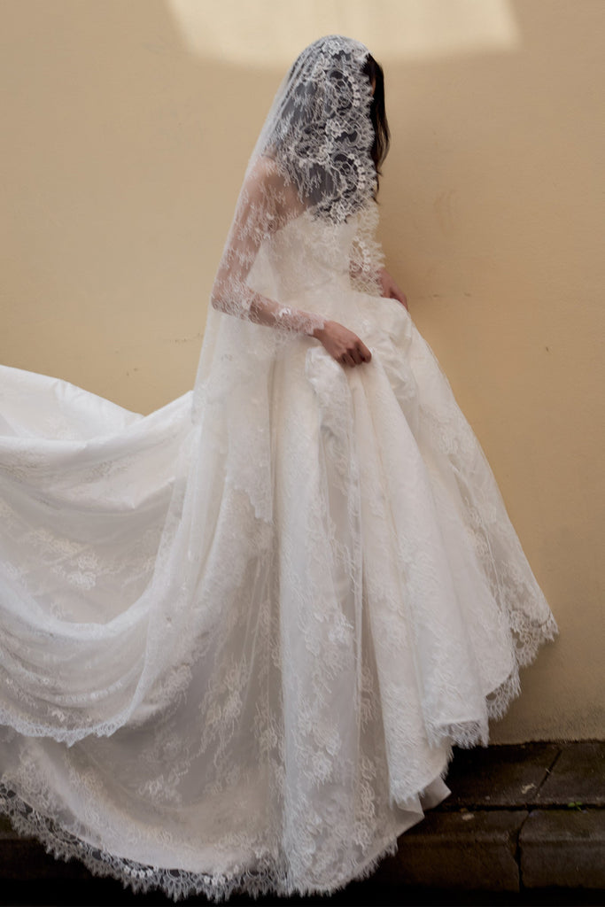 A woman wearing a voluminous white lace wedding gown with a long sheer veil, photographed from behind against a plain beige wall.