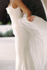 A woman wearing a white sleeveless wedding dress with a fitted bodice and a flowing sheer tulle overskirt, photographed in soft natural light.