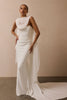 A model wearing a floor-length white draped gown with a high draped neckline, ruched bodice, and a long flowing train against a warm beige studio wall.