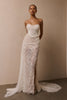 A woman modeling a strapless white lace wedding gown with a fitted bodice, thigh-high slit, and long sheer train against a plain beige wall_XS_