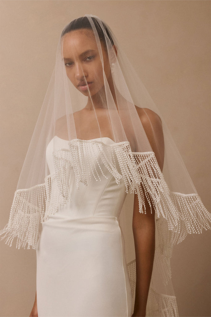 A woman wearing a long sheer white bridal veil with beaded fringe draped over her shoulders and a white strapless gown against a warm beige studio background.