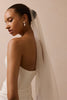 A woman in a white strapless wedding gown shown from the back, wearing a long sheer bridal veil and pearl drop earrings against a soft beige studio background.