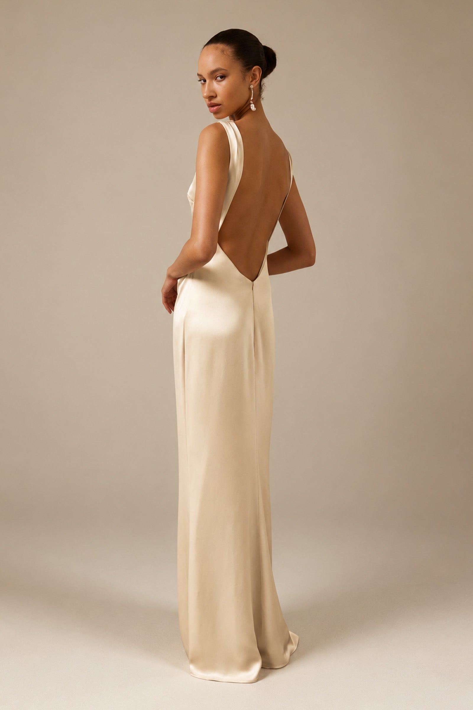 A woman modeling a floor-length cream satin gown from the back, showing a deep open back and draped neckline, set against a solid warm beige studio background_XS_