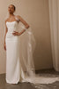A woman modeling a strapless white wedding gown with a long sheer veil featuring floral embroidery, posed against a neutral beige wall.