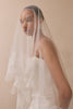 A woman wearing a long white bridal veil with lace floral applique and a white wedding dress, photographed against a soft beige studio background.