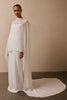 A model wearing a flowing white high-low dress with a sheer draped capelet overlay, standing against a neutral beige wall.