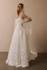 A woman modeling a white lace bridal gown with a long sheer veil and fringe detail, standing against a plain beige studio wall.