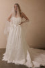 A woman modeling a strapless white lace wedding gown with a long sheer veil, standing against a plain beige wall.