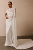 A model wearing a floor-length white gown with a high mock neck, draped cape-like sleeves, and a long train, standing against a plain beige wall.