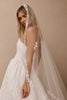 A woman wearing a white wedding dress and a long sheer tulle bridal veil with floral lace applique, posed against a warm beige studio background.
