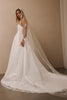 A woman modeling a white strapless wedding gown with a long sheer veil and floral lace appliques, posed in a minimalist indoor setting with a warm beige wall.