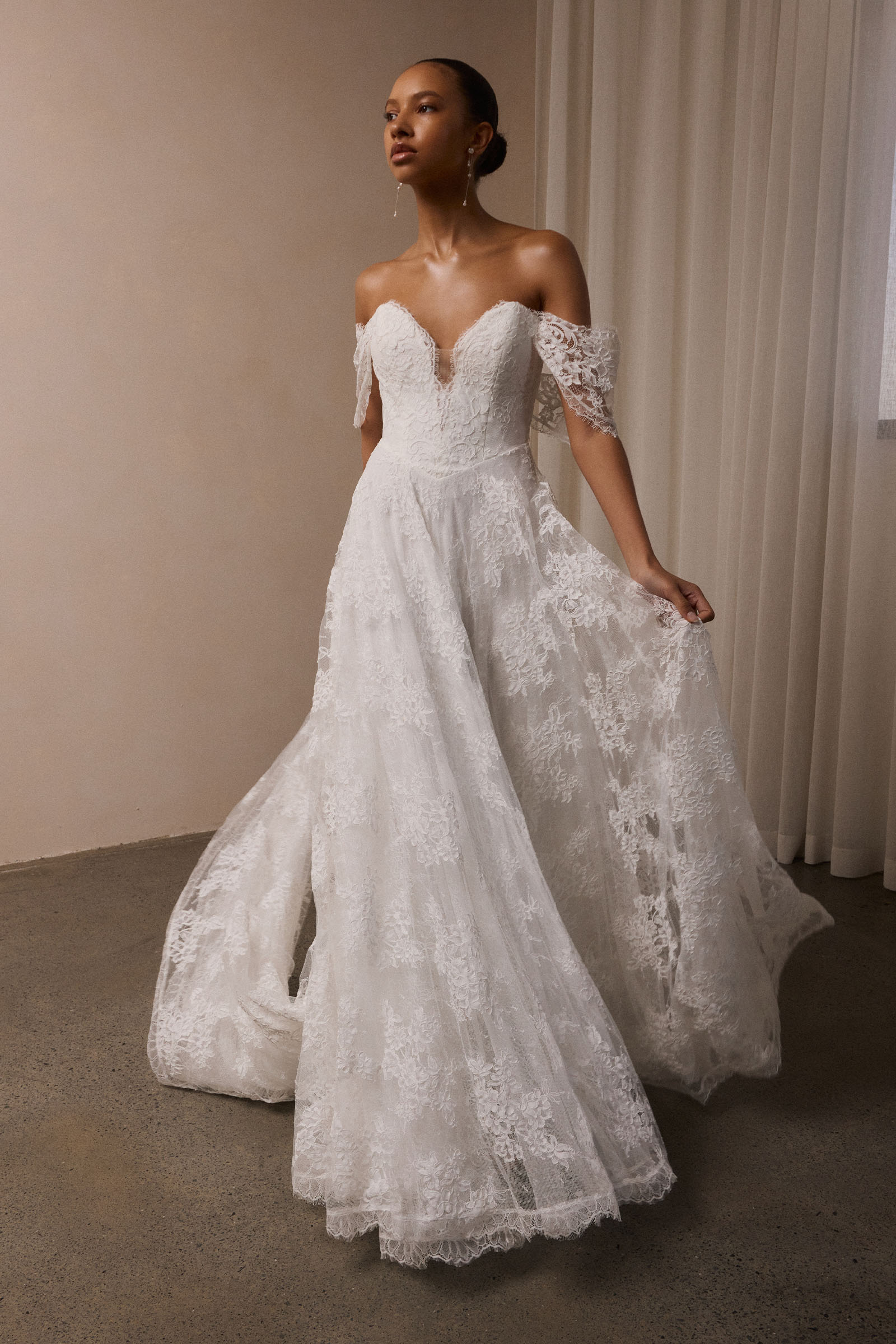 A woman wearing a white off-the-shoulder lace wedding gown with a deep V neckline and a full layered skirt_XS_