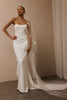 A woman modeling a strapless white mermaid-style wedding gown with a long sheer veil featuring floral lace applique, standing in a softly lit indoor setting with a beige wall and floor.