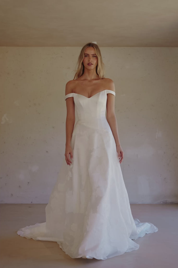Video of a woman wearing an off-the-shoulder white wedding dress with a fitted bodice and flowing skirt